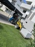 Gelber Radlader transportiert einen Stapel Gehwegplatten auf einer Palette vor einem modernen Wohnhaus.