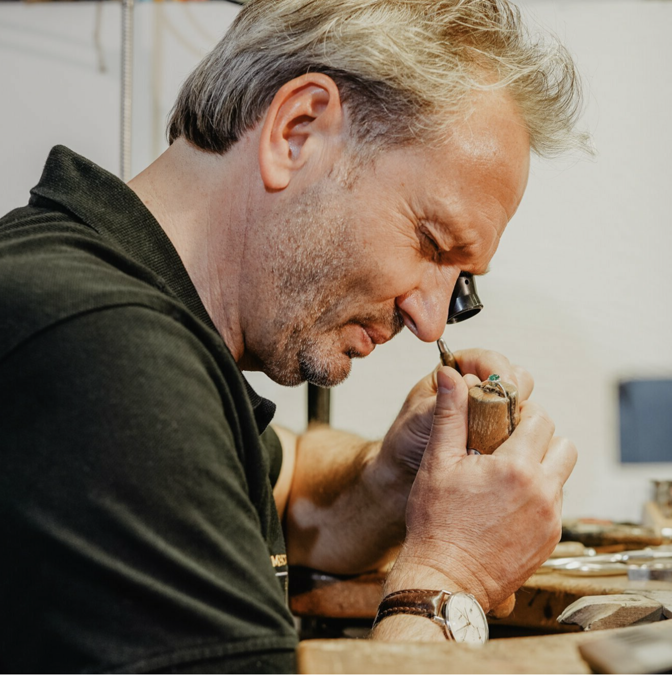 Goldschmied mit Uhrmacherlupe bearbeitet einen Ring mit grünem Edelstein an einem hölzernen Werktisch.