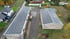 Zwei große Hallen mit Solarmodulen auf den Dächern, ein LKW dazwischen, daneben Häuser und Bäume.