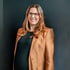 Lächelnde Frau mit Brille, langen braunen Haaren und kupferfarbenem Blazer vor einer dunklen Wand.
