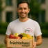 Ein lächelnder Mann in weißem T-Shirt mit "früchtehaus Stuttgart"-Logo hält eine Holzkiste voller frischer Früchte.