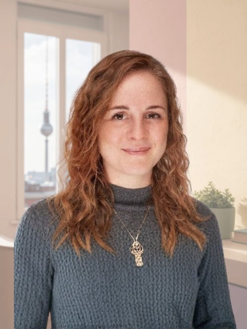 Lächelnde Frau mit langen rotbraunen Locken und Halskette vor einem Fenster mit Blick auf den Berliner Fernsehturm.