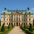 Schloss mit hellen Mauern, grünem Kupferdach und zwei Türmen, davor ein Kiesweg und getrimmte Hecken.