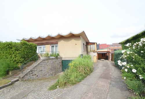 Beigefarbenes Haus mit wellenförmigem Dach, Steinmauer, Zufahrt, grünen Büschen und weißen Blumen.