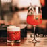 Zwei rote Cocktails, einer in einem niedrigen Glas mit Schaum, der andere in einem Weinglas mit Beeren und Strohhalm.