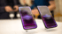 
Das Bild zeigt zwei iPhones, die auf einem weißen Tisch stehen. Die Bildschirme beider Geräte sind beleuchtet und zeigen den Text "Hello, Apple Intelligence." in der Mitte. Die Hintergrundbeleuchtung der Bildschirme ist eine Mischung aus Lila- und Rosatönen, die nach unten hin dunkler werden. Der Fokus liegt auf den beiden Handys, während der Hintergrund unscharf ist. Im Hintergrund sind undeutliche Umrisse von Personen zu erkennen.