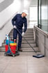 Ein Hausmeister in blauer Uniform und weißer Kappe wischt einen gefliesten Boden neben einer Treppe mit Reinigungswagen.