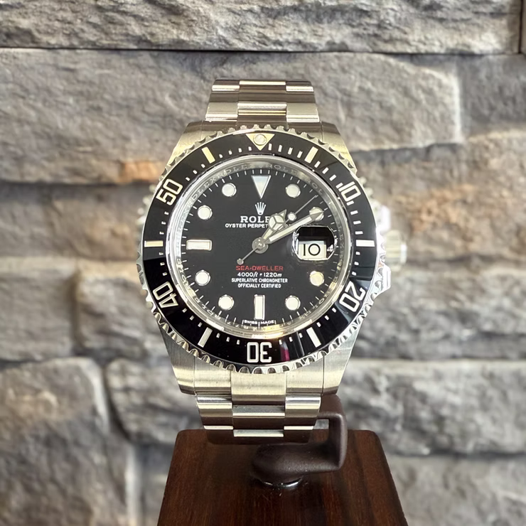 Eine silberne Rolex Sea-Dweller Uhr mit schwarzem Zifferblatt und Datumsanzeige auf einem Holzständer vor einer Steinwand.