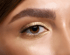 Nahaufnahme eines Auges mit brauner Iris und goldenem Lidschatten, umrahmt von einer vollen Augenbraue.