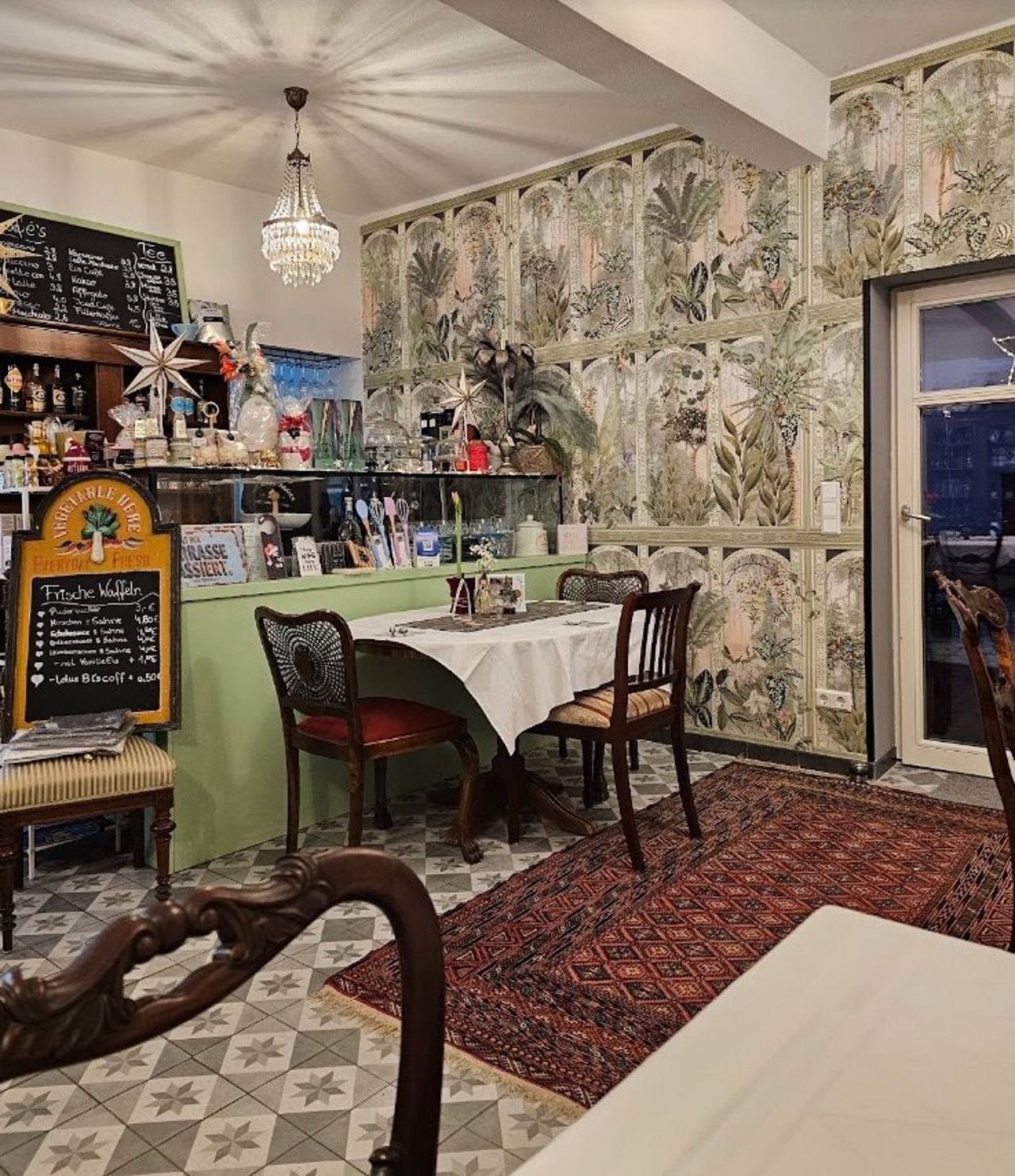Gastraum des Café Löwenmäulchen mit Palmentapete, Kronleuchter, mintgrüner Theke, Zementfliesen und antiken Holzstühlen