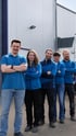 Fünf lächelnde Mitarbeiter in blauer Arbeitskleidung stehen mit verschränkten Armen vor einem Firmengebäude.