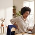 A woman sits at a desk using a laptop while holding her lower back with one hand.