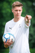 Ein junger Mann im weißen T-Shirt mit Puma-Logo hält einen Fußball und zeigt mit dem Finger nach vorne.