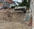 Baustelle mit ausgehobener Grube, Schutt, Bagger und einem großen, liegenden, verrosteten Metalltank.