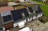 Haus mit Solarmodulen auf dem Dach, zwei Personen arbeiten vor dem Haus, Anhänger parkt auf dem Hof.