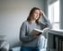 A woman in a grey sweater holds an open book and leans against a bookshelf with a concerned expression.