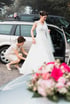 Eine Frau in einem weißen Hochzeitskleid steigt aus einem Auto, während eine andere Frau ihr Kleid richtet.