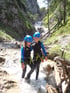 Zwei Kinder in Neoprenanzügen und Helmen stehen in einem Bach vor einem Wasserfall.