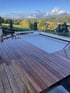 Ein geschlossener Pool mit weißer Lamellenabdeckung auf einer Holzterrasse vor einer hügeligen Landschaft.