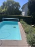 Ein Poolroboter reinigt ein hellblaues Schwimmbecken im Garten neben einer Hecke und einem weißen Haus.