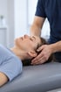 Ein Physiotherapeut führt eine manuelle Therapie am Kopf einer liegenden Frau durch.