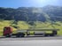 Roter Lastwagen transportiert lange Betonplatte auf Anhänger vor grünen Bergen.