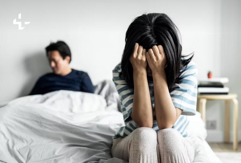 Eine Frau sitzt auf dem Bett und hält sich die Hände vors Gesicht, ihr Partner im Hintergrund