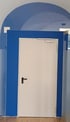 Weiße Tür mit schwarzem Griff in einem blauen Rahmen mit gewölbter blauer Decke.