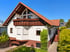Weißes Einfamilienhaus mit rotem Ziegeldach und Holzbalkon, umgeben von grünen Büschen unter blauem Himmel.