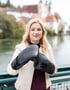 Blonde Frau in beigefarbener Jacke hält schwarze Boxhandschuhe vor einem Fluss und einer Stadt.