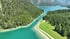 Türkisfarbenes Wasser eines Bergsees fließt durch einen von grünen Nadelwäldern gesäumten Kanal, über den eine Brücke führt.