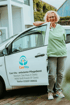 Eine lächelnde Frau in hellgrüner Arbeitskleidung lehnt an einem weißen Auto mit „CareVita – ambulanter Pflegedienst“-Aufschrift.