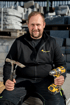 Ein lächelnder Handwerker hält einen Hammer und einen gelben Akkuschrauber vor einem Lager mit Steinplatten.