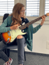 A young person with glasses and ripped jeans sits, playing an electric guitar.