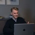 Ein junger Mann mit blonden Haaren und Bart lächelt und blickt auf einen geöffneten Laptop.