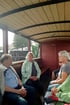 Mehrere Personen sitzen in einem offenen Waggon mit Holzdach und roten Seitenwänden, schauen zur Seite.