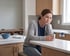 A woman sits at a kitchen counter holding a mug with her chin resting on her hand, looking away with a somber expression.