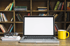 Laptop, Tasse, Brille und Notizbüchern vor einem Bücherregal auf einem Holztisch.