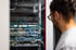 Mann mit Brille betrachtet Serverrack mit Netzwerkgeräten und vielen farbigen Kabeln.