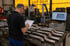 Ein Mitarbeiter prüft Metallrohre in einer Werkstatt mit Klemmbrett, Faro-Messarm und CAD-Modell am Monitor.