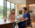 A male healthcare professional in blue scrubs shows a digital tablet with data charts to a smiling woman at a wooden table.