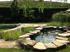 Teich mit Steinumrandung, Seerosen und einem kleinen Springbrunnen in einem Garten mit Hecke und Bäumen.