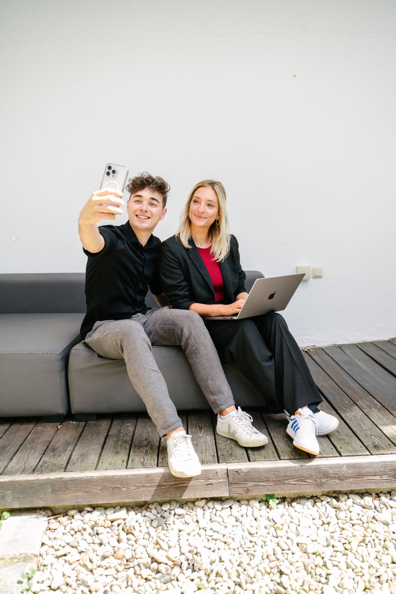 Ein junger Mann macht ein Selfie mit einer Frau, die an einem Laptop arbeitet, auf einer grauen Couch im Freien.
