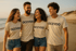 Vier lachende junge Erwachsene in beigen T-Shirts mit dem Text "OCEVOO" gehen am Strand.