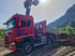 Roter Scania-LKW mit Kran und Kippmulde auf einer Baustelle mit Erdhügeln und Bergen im Hintergrund.