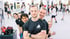 Ein lächelnder Boxtrainer mit verschränkten Armen steht vor seiner Gruppe in einer hellen Kampfsport-Trainingshalle.