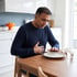 A man in a blue sweater sits at a kitchen table, holding his stomach in discomfort after finishing a meal.