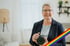Eine lächelnde Frau mit kurzem grauen Haar und Brille hält eine schwarze Kamera, mit Regenbogen-Streifen.