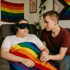 Eine Person mit Schlafmaske und Kopfhörern sitzt unter einer Regenbogenfahndecke, während eine andere Person ihre Hand hält.
