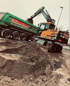 Ein Hyundai Mobilbagger belädt auf einer Baustelle einen grünen LKW des HAAK Containerdienstes mit Erde.